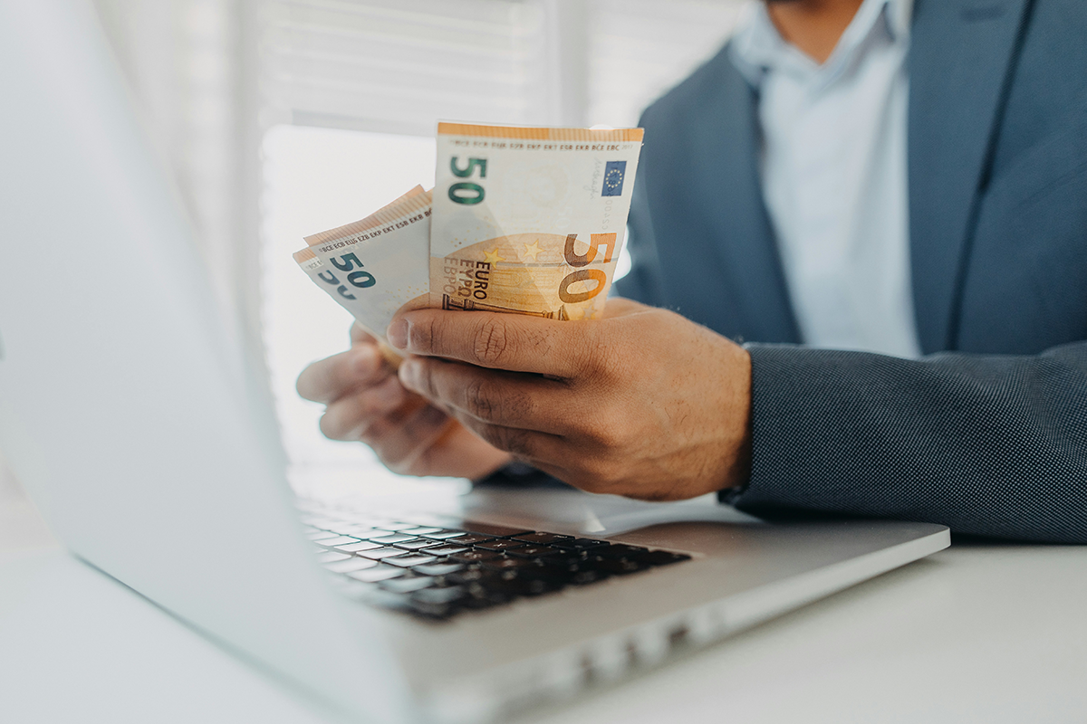 A person counting euro banknotes at a laptop